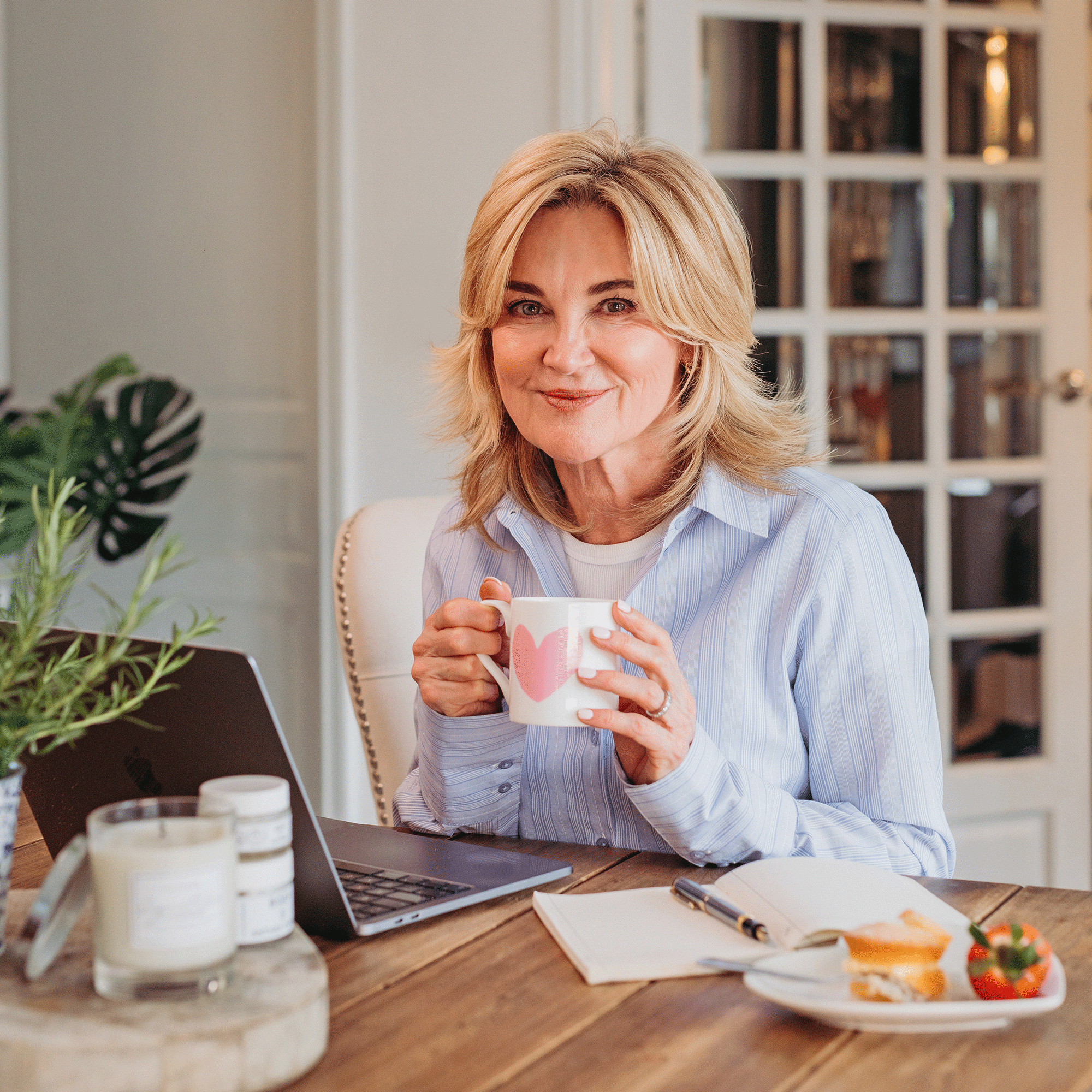 Anthea Turner wearing blue striped shirt in a kitchen sitting at a table with laptop and mug