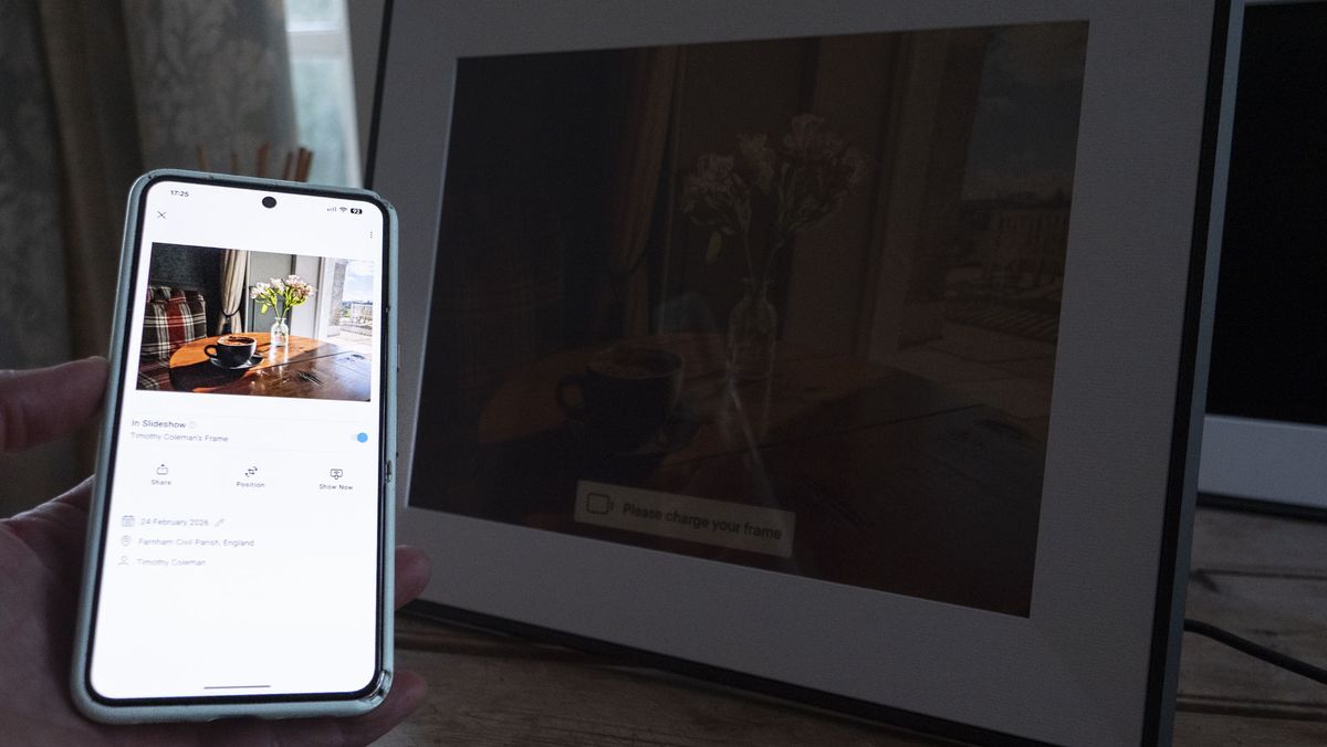 A phone being held, displaying the Aura companion app for digital photo frames, in front of the The Aura Ink 'E Ink' digital photo frame on a sideboard