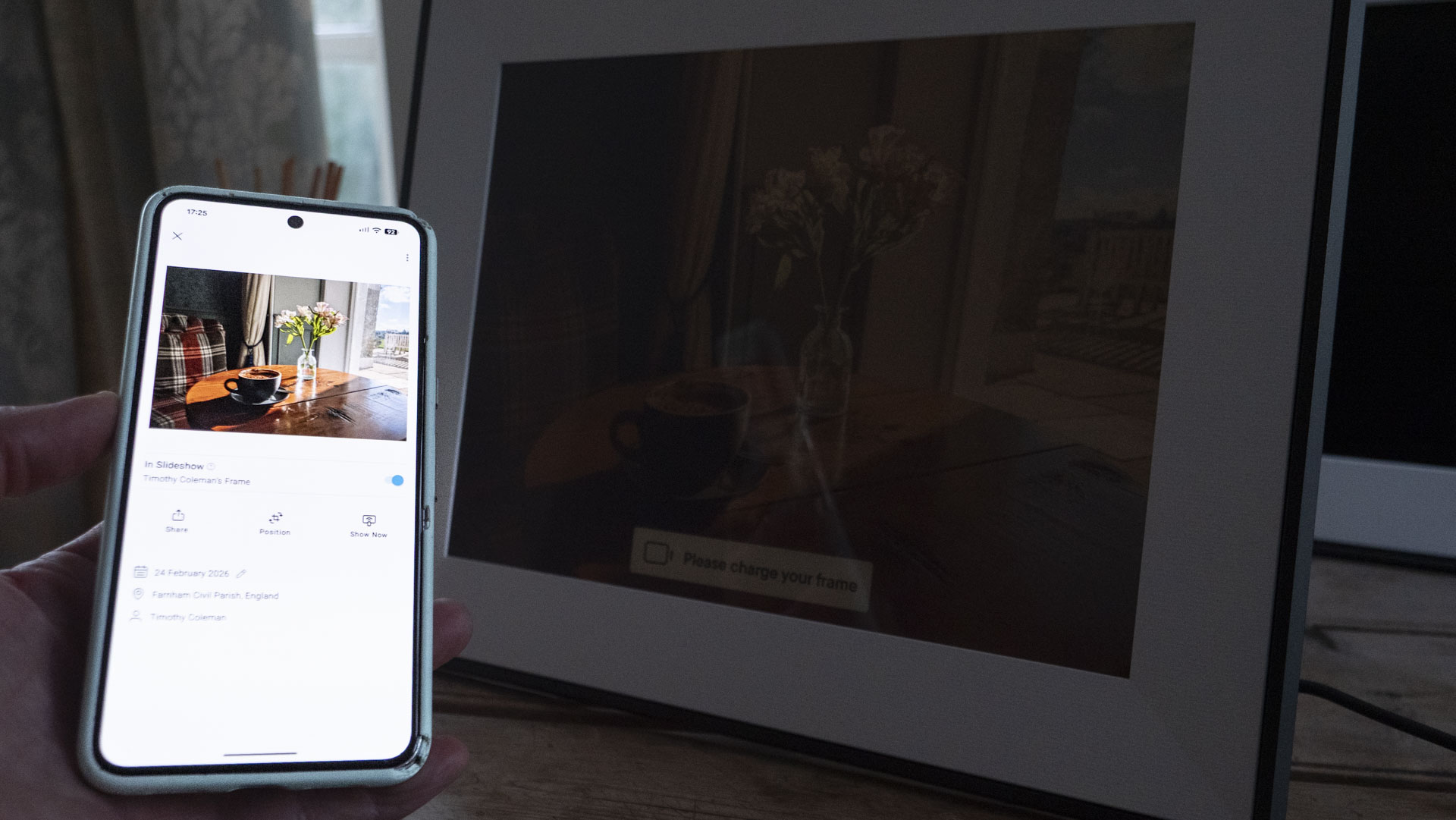 A phone being held, displaying the Aura companion app for digital photo frames, in front of the The Aura Ink 'E Ink' digital photo frame on a sideboard