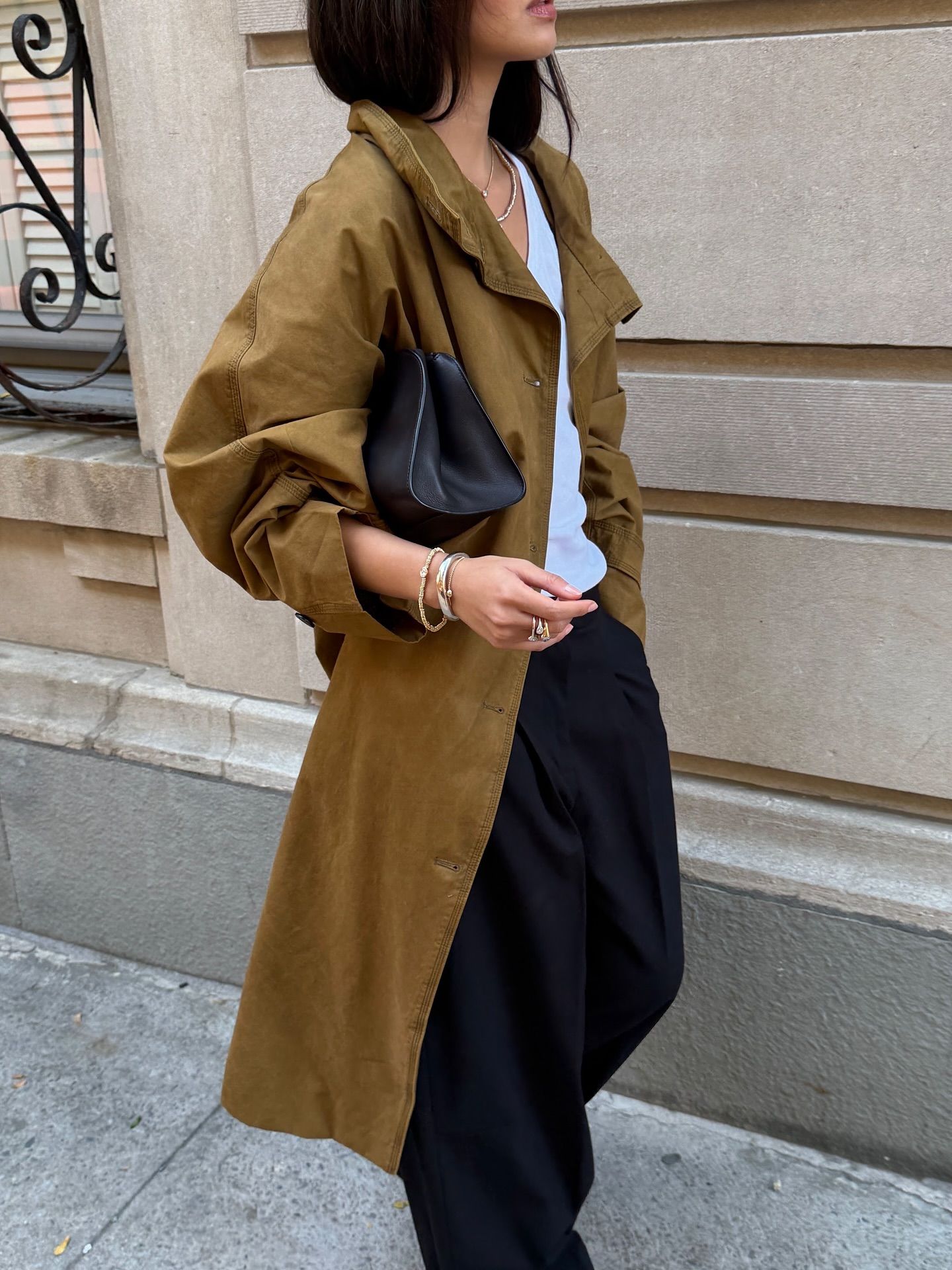 An image of an influencer in New York wearing a dark beige trench coat with black trousers and a white top, also wearing stacks of bracelets on her wrist and a ring stack on her finger
