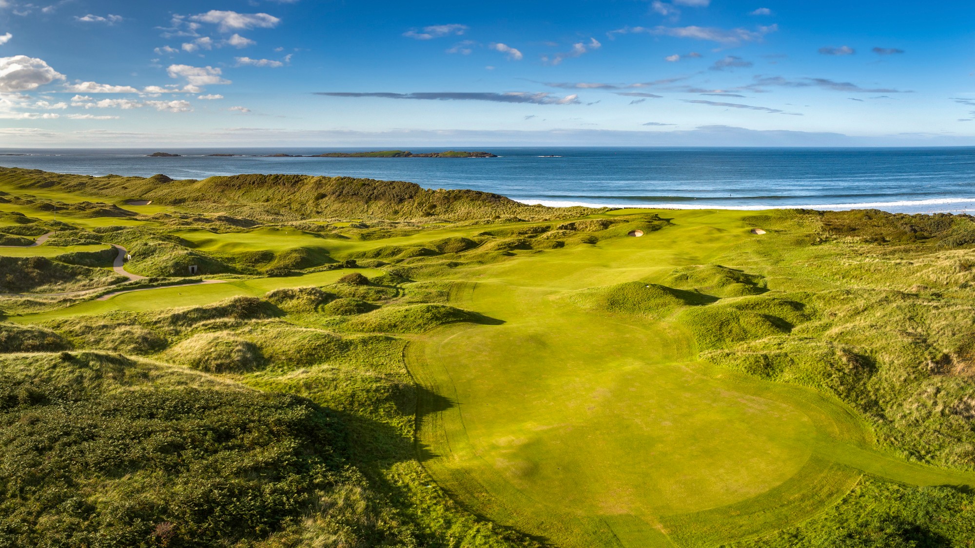 The 5th hole at the Royal Portrush Golf Course in Ireland. The aerial view shows a lush greenway with the ocean in the background.