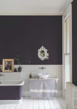 A plum colored bathroom with white painted floorboards, a white mirror and artwork on a shelf