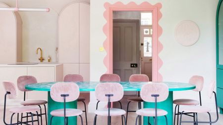 A white dining room and open kitchen with a colorful peachy gradient frame around the door way. A deep turquoise dining table is surrounded by pale pink chairs with black metal accents