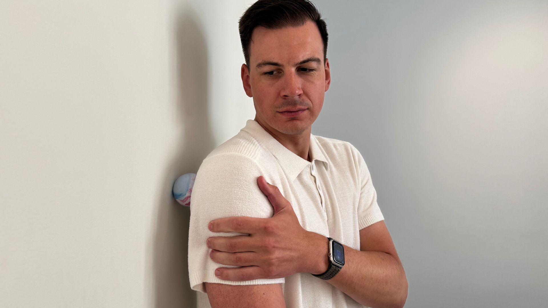 Man in white polo shirt pressing his shoulder blade into a ball which is pressing into a wall
