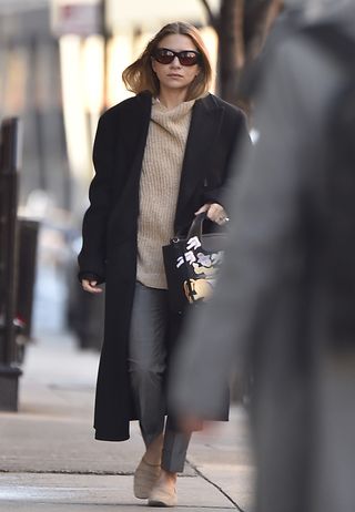 Ashley Olsen walking in New York City wearing oversize sunglasses, an oversize black coat, a ribbed neutral mockneck turtleneck sweater, gray pants, and tan suede loafers.