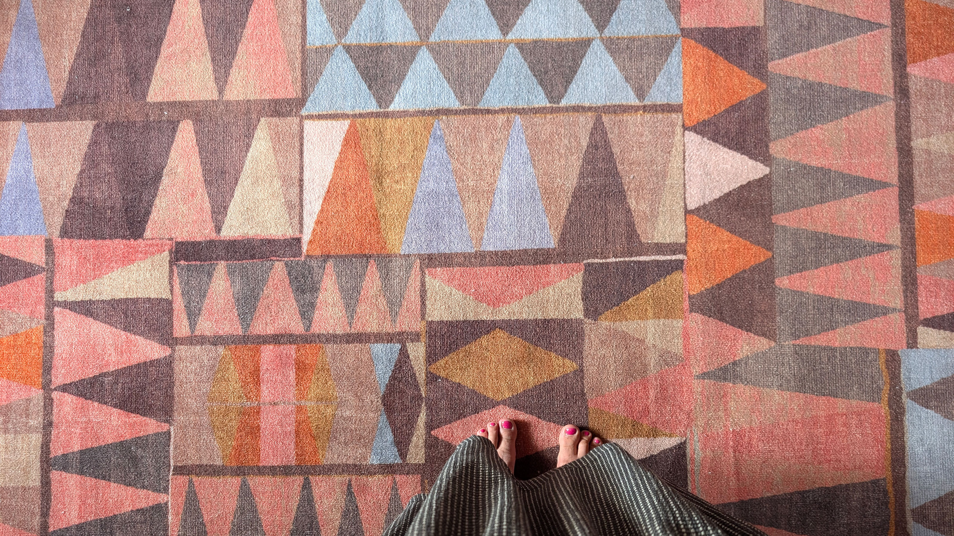 Point of view of woman looking down onto feet while standing on rug with abstract pattern in oranges, browns, pinks and blues