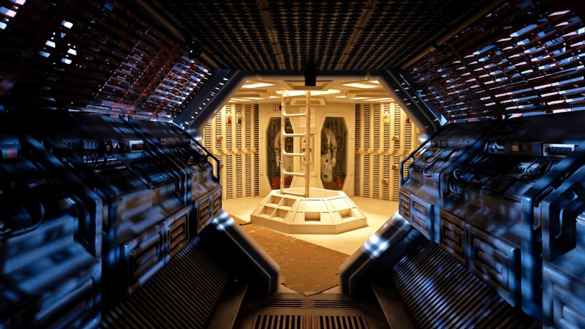 a view down dimly corridor lined with pipes and ducts leading into a bright, yellow-tinged room containing a ladder and spacesuits hung on a rack