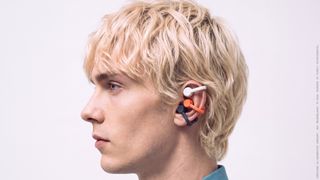A man with blonde hair in profile, wearing three pairs of open earbuds in white, black and coral, on white background