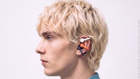A man with blonde hair in profile, wearing three pairs of open earbuds in white, black and coral, on white background