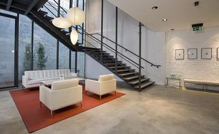 Open ground floor space, neutral floor, square textured red rug with a cream sofa and two armchairs on top, black stairwell with black handrails, white brick walls, black framed pictures, large glass front with black frame doorway and external shrubs in view against a brick wall, white ceiling with spotlights, white round and oval shaped ceiling lights, small glass side table and white wall bench