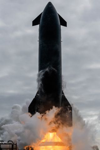 a tall pointy rocket with a black belly fires its engines on a test stand with smoke billowing to the left.