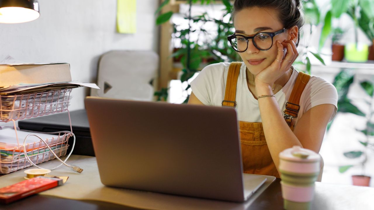 Young woman looking at an open laptop in front of her with office supplies on the desk around her