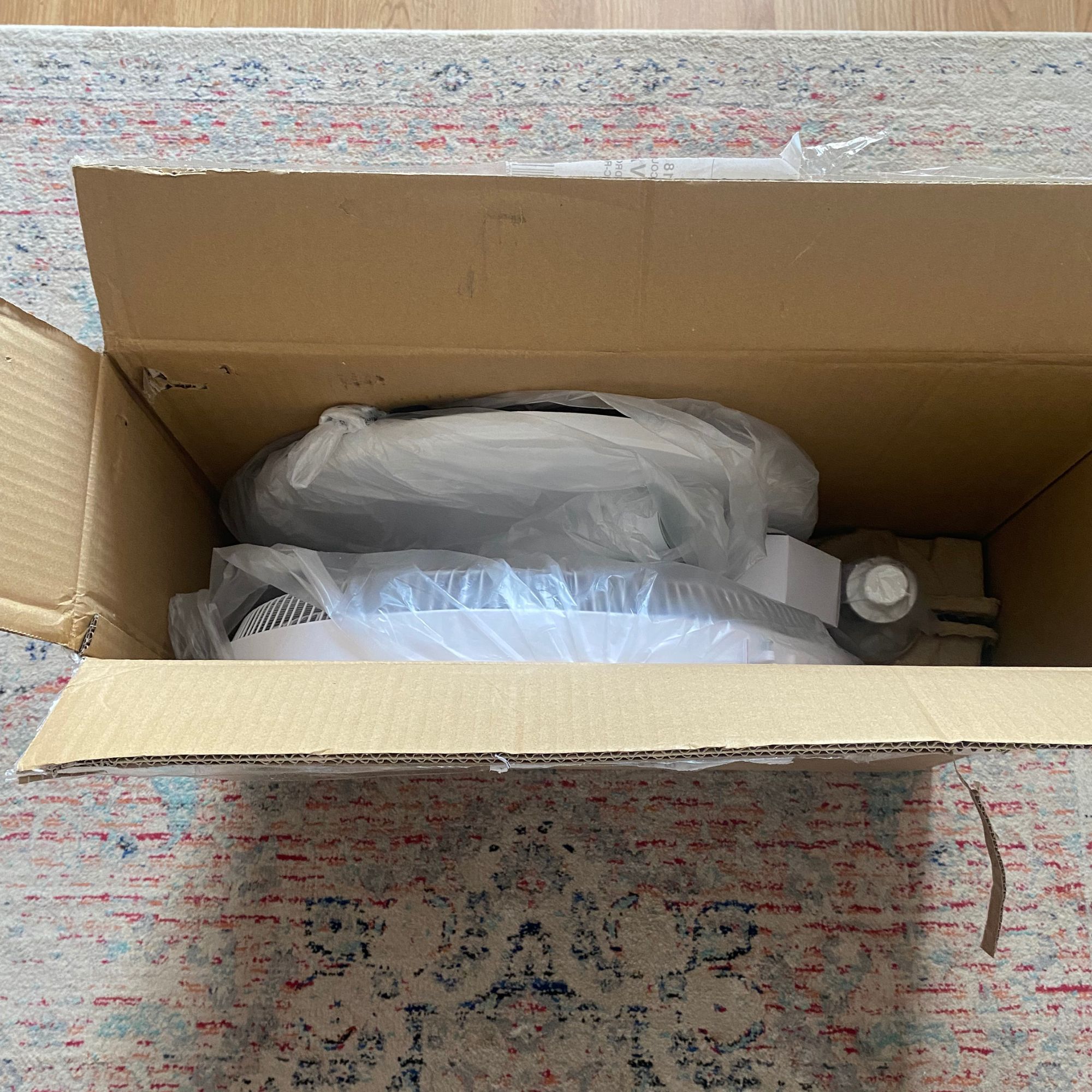 An open cradboard box with the Vonhaus 16'' pedestal fan inside, sitting on a patterned rug