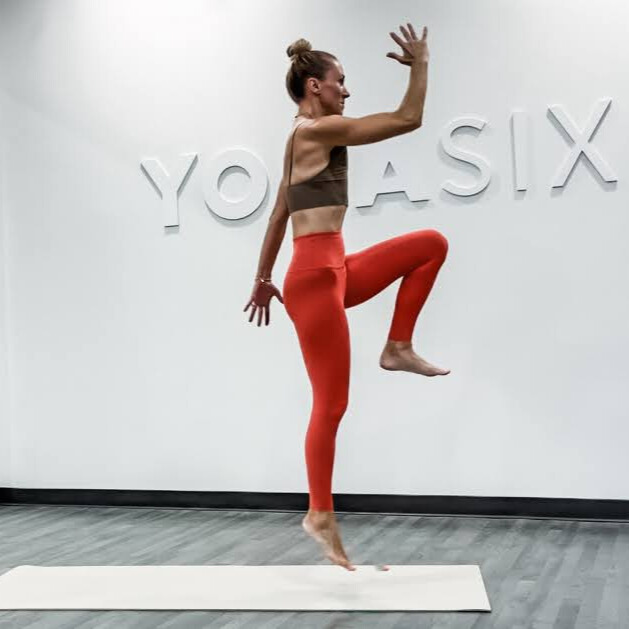 Woman demonstrates yoga pose in yoga studio with words Yoga Six embossed on wall behind her