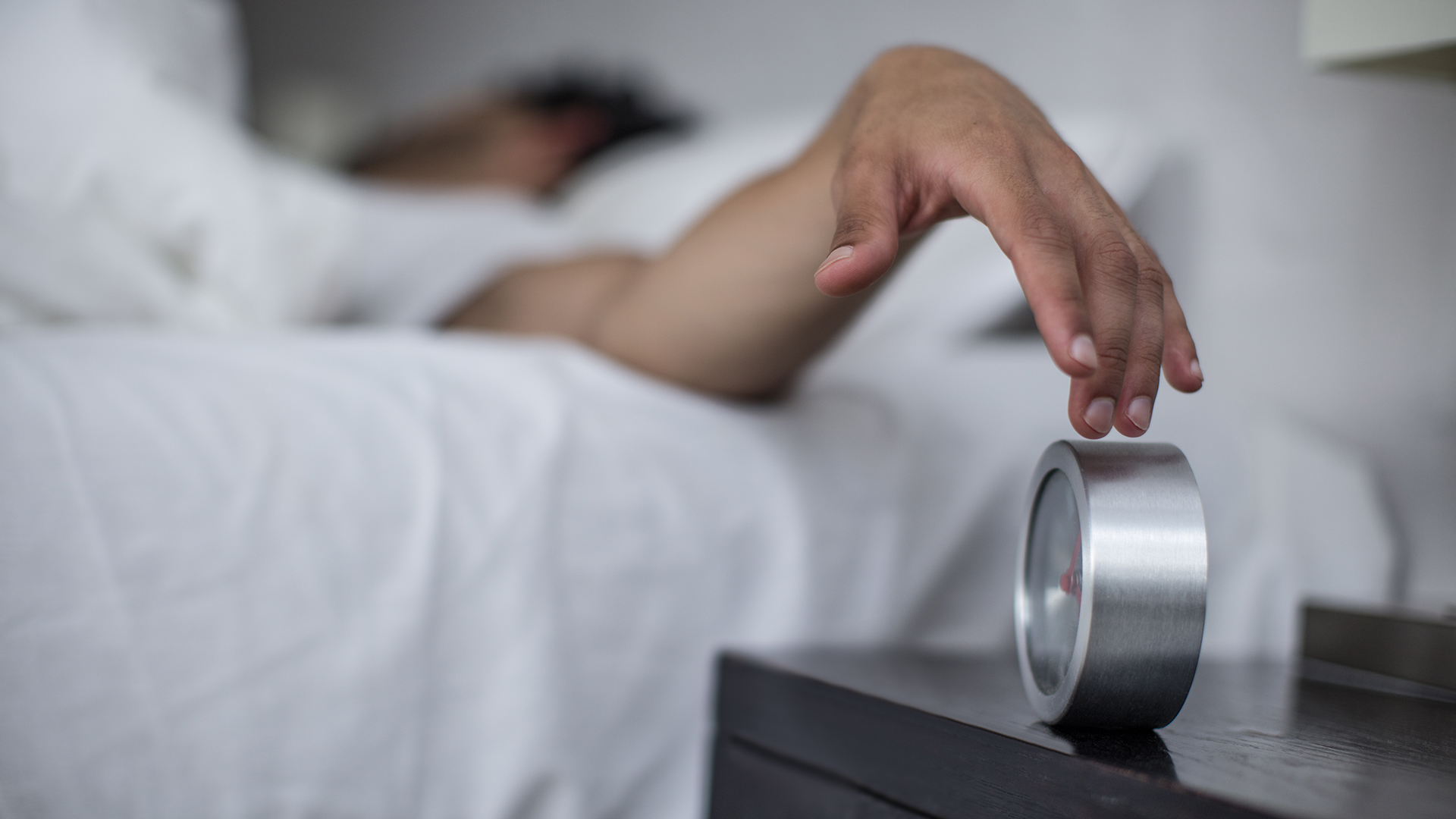 A man reaches out of bed to turn off his alarm clock