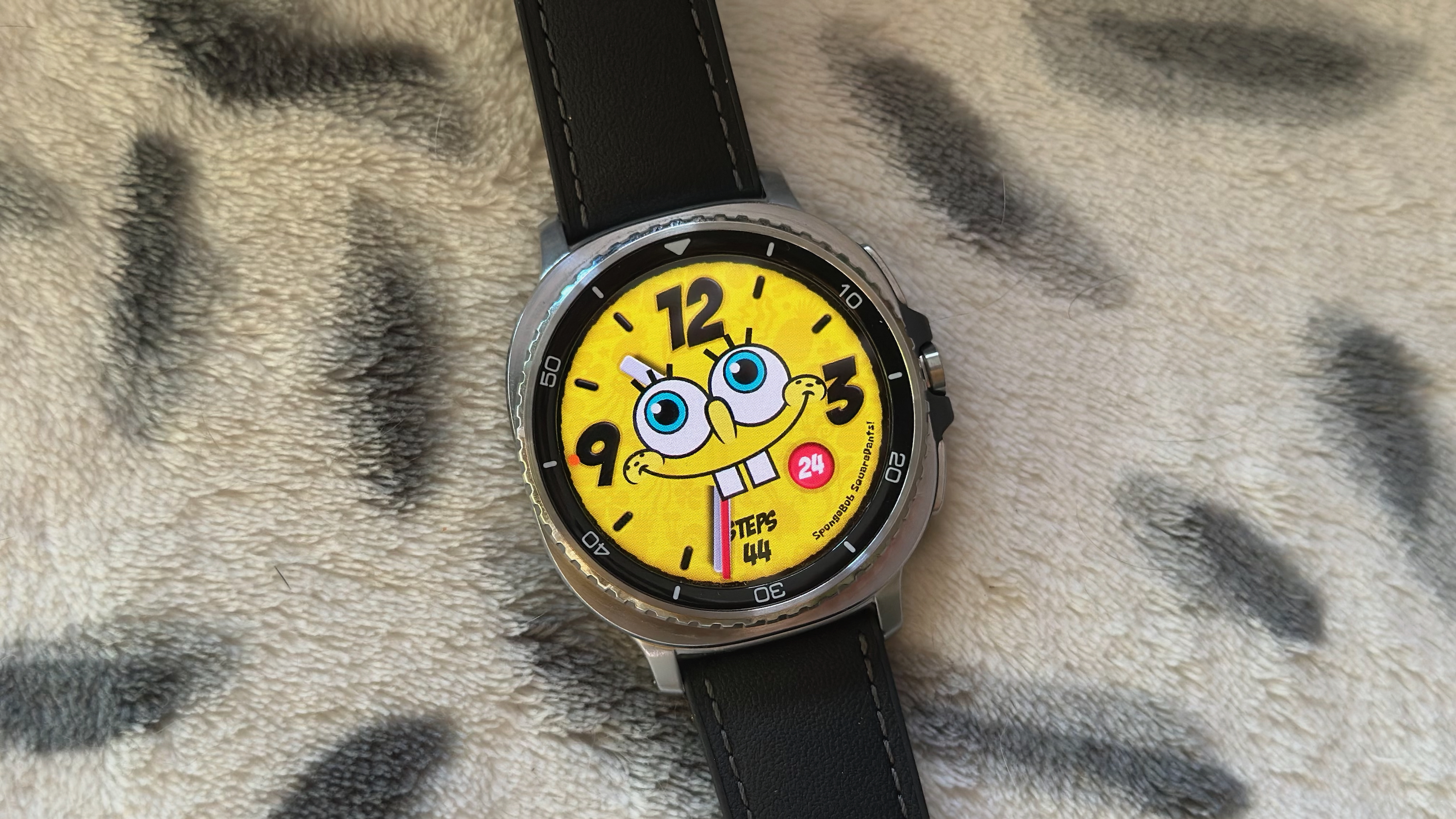The Samsung Galaxy Watch 8 Classic resting on a blanket showing a custom Facer watch face of Spongebob Squarepants.