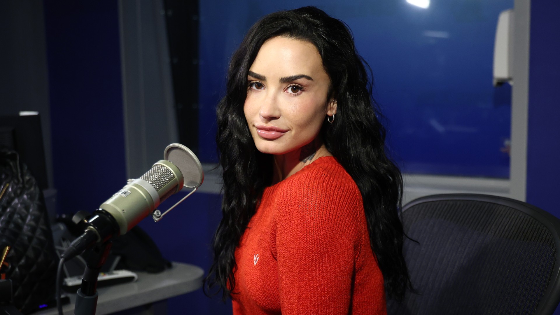 a white woman with dark hair wearing a bright red sweater and posing in front of a microphone in a studio