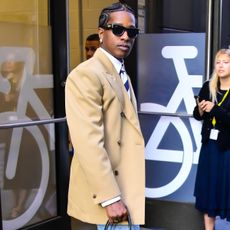 Asap rocky wears a beige blazer, stripe tie, sunglasses, and celine's smile luggage bag.