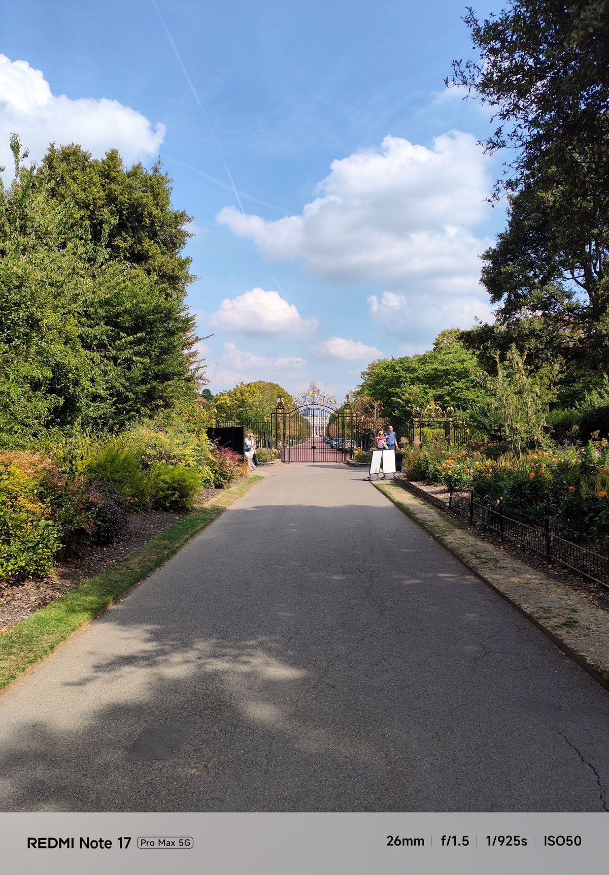 Tree-lined pathway through a landscaped garden leading toward ornate gates beneath a bright blue sky.