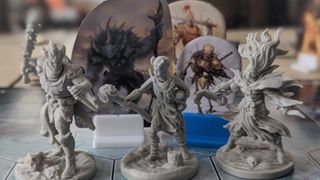 Three Frosthaven miniatures are lined up on a board in front of cardboard monster standees