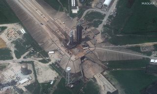 A high-altitude aerial view of SpaceX's triple-booster Falcon Heavy rocket can be seen standing in the center of the wide landscaped launch complex. Roads, pipes and other infrastructure extend outward from the rocket and adjacent launch tower.