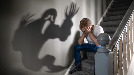 Man sitting on a staircase with his head in his hands, with the shadow of a ghost haunting him on the wall next to him