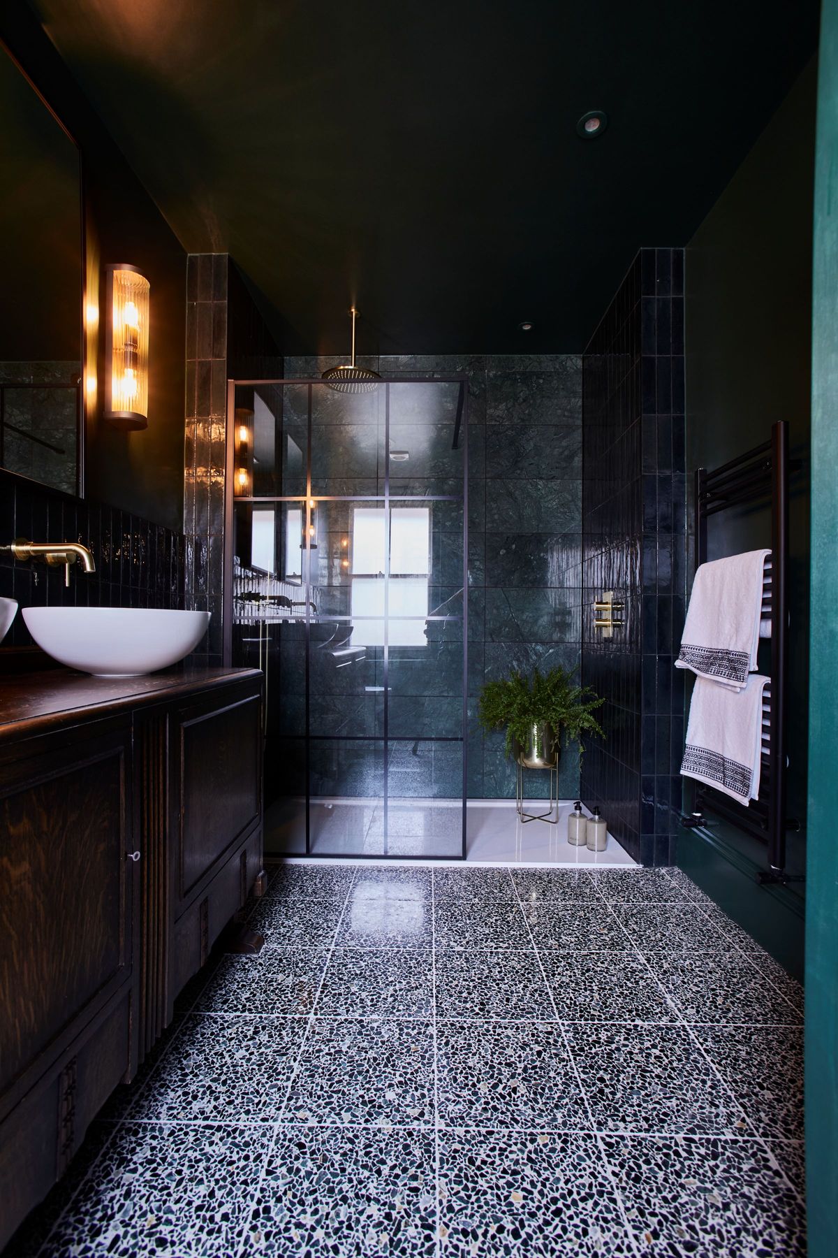 A completely black tiled bathroom with a walk in shower with a white sink and mirror