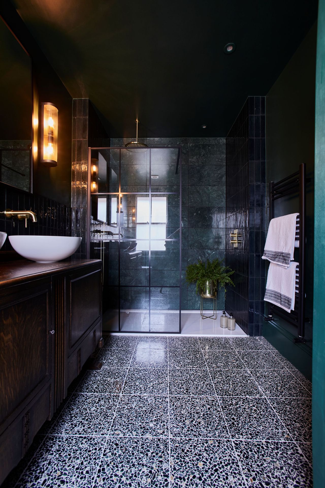 A completely black tiled bathroom with a walk in shower with a white sink and mirror