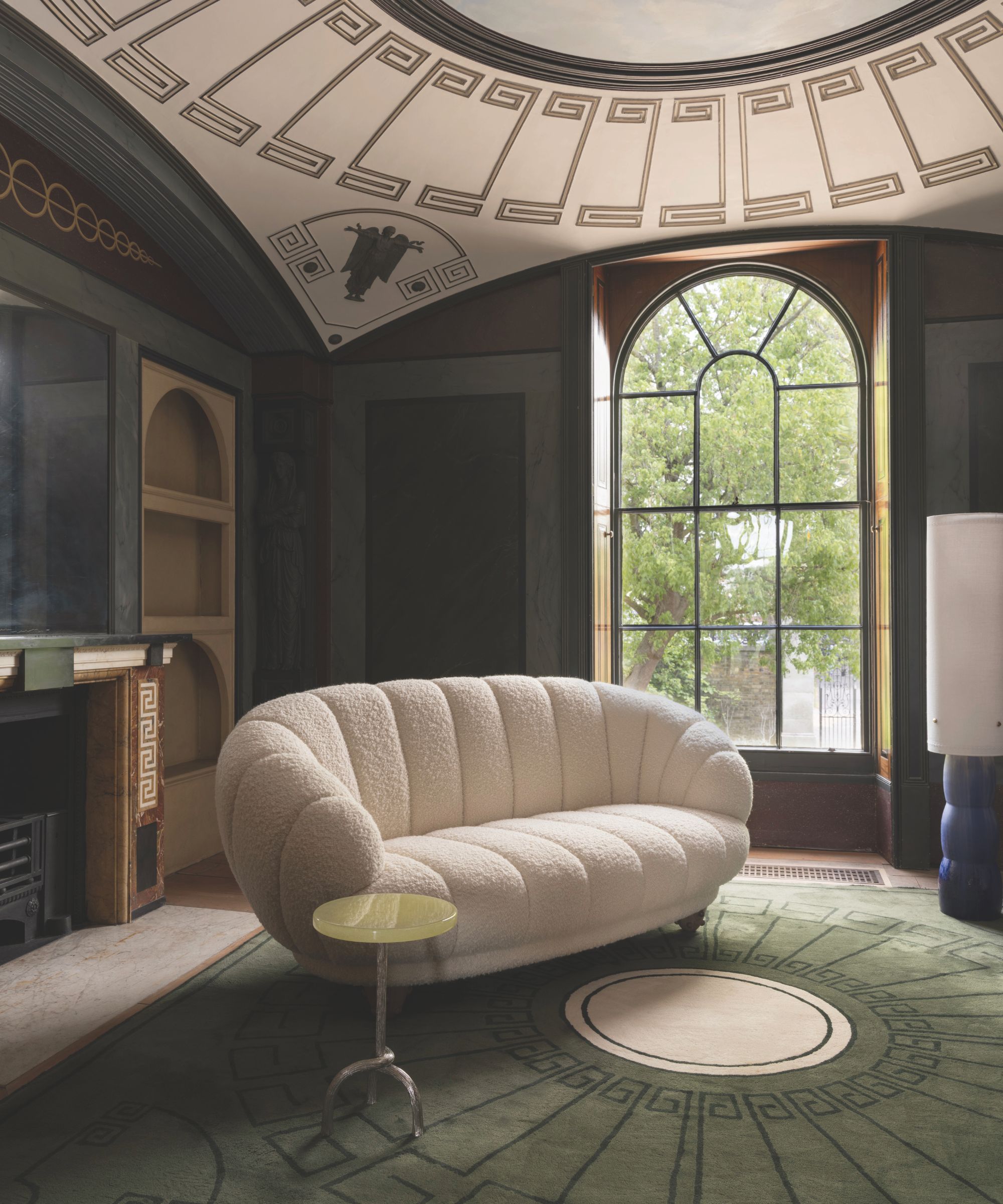 A dark blue room with a large green patterned rug in the middle. On the rug is a white boucle sofa and a small circular table. One wall has a large window.