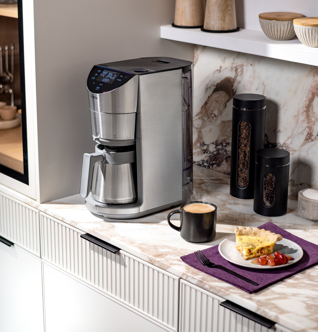 Image of a silver AI coffee machine on a marble kitchen counter. There is a cup of coffee and a plate of breakfast next to the machine.