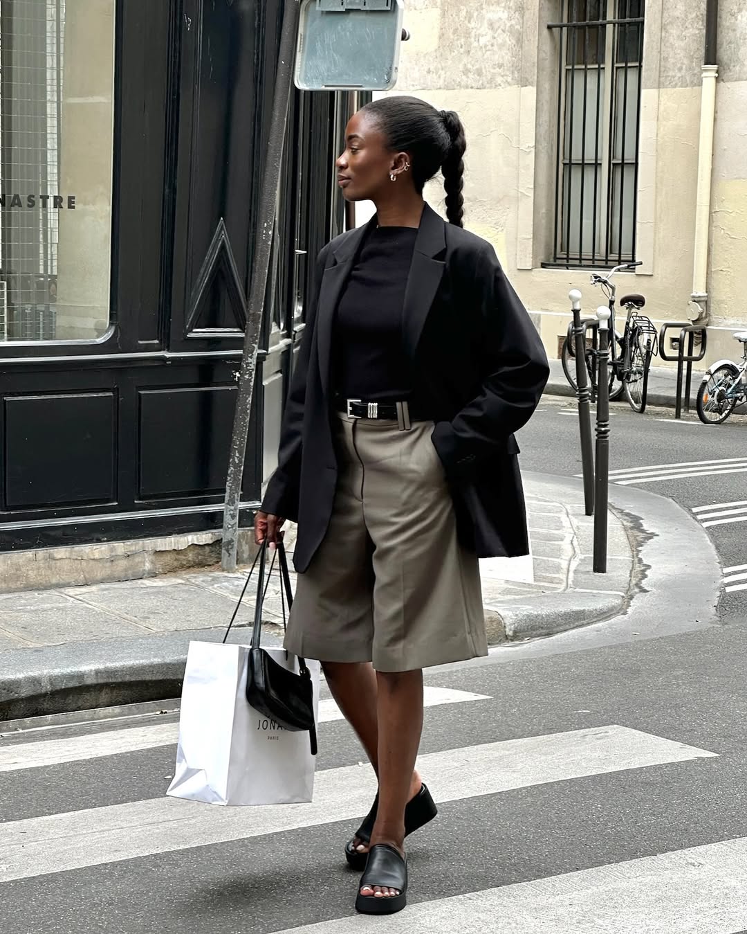 @_livmadeline wears a black linen blazer, long grey-green shorts, black slides and a shoulder bag