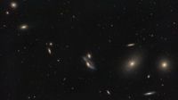 A string of galaxies shines in a deep space photo in a black sky dotted with stars.