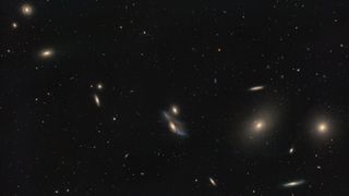 A string of galaxies shines in a deep space photo in a black sky dotted with stars.
