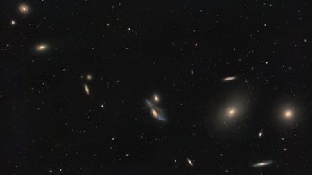 A string of galaxies shines in a deep space photo in a black sky dotted with stars.