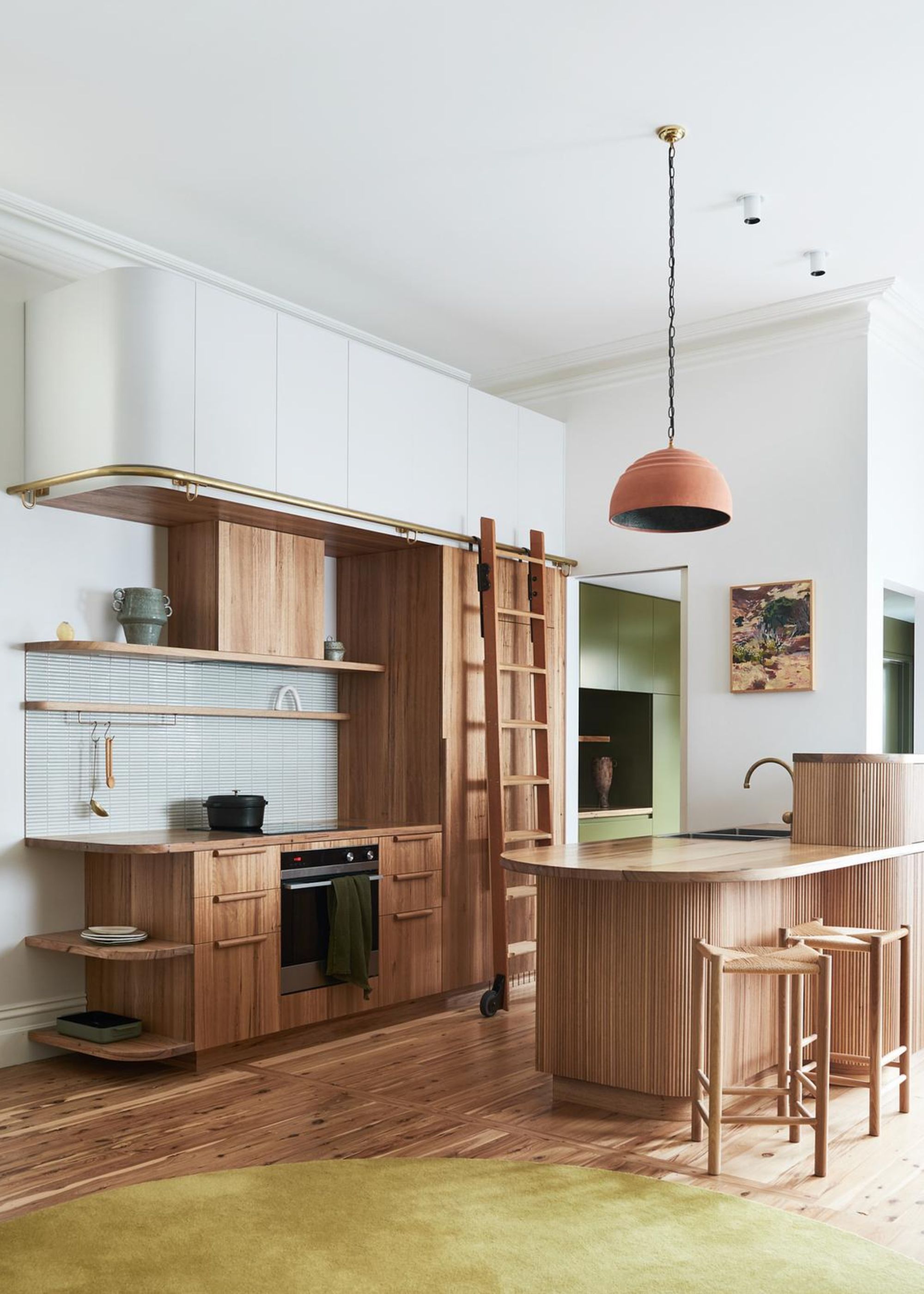 A kitchen with a curved island/seating area, as well as curved kitchen shelving with a view of the oven and stove top. There is also a ladder in the kitchen with gives access to a group of white colored cabinets.