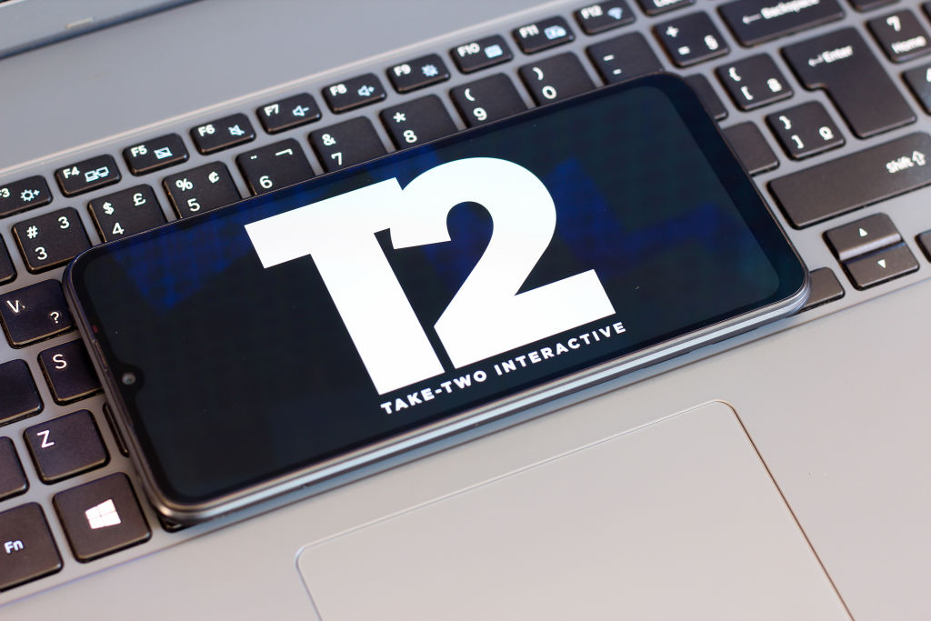 The Take-Two Interactive (T2) logo is seen displayed on a smartphone screen next to a laptop keyboard.