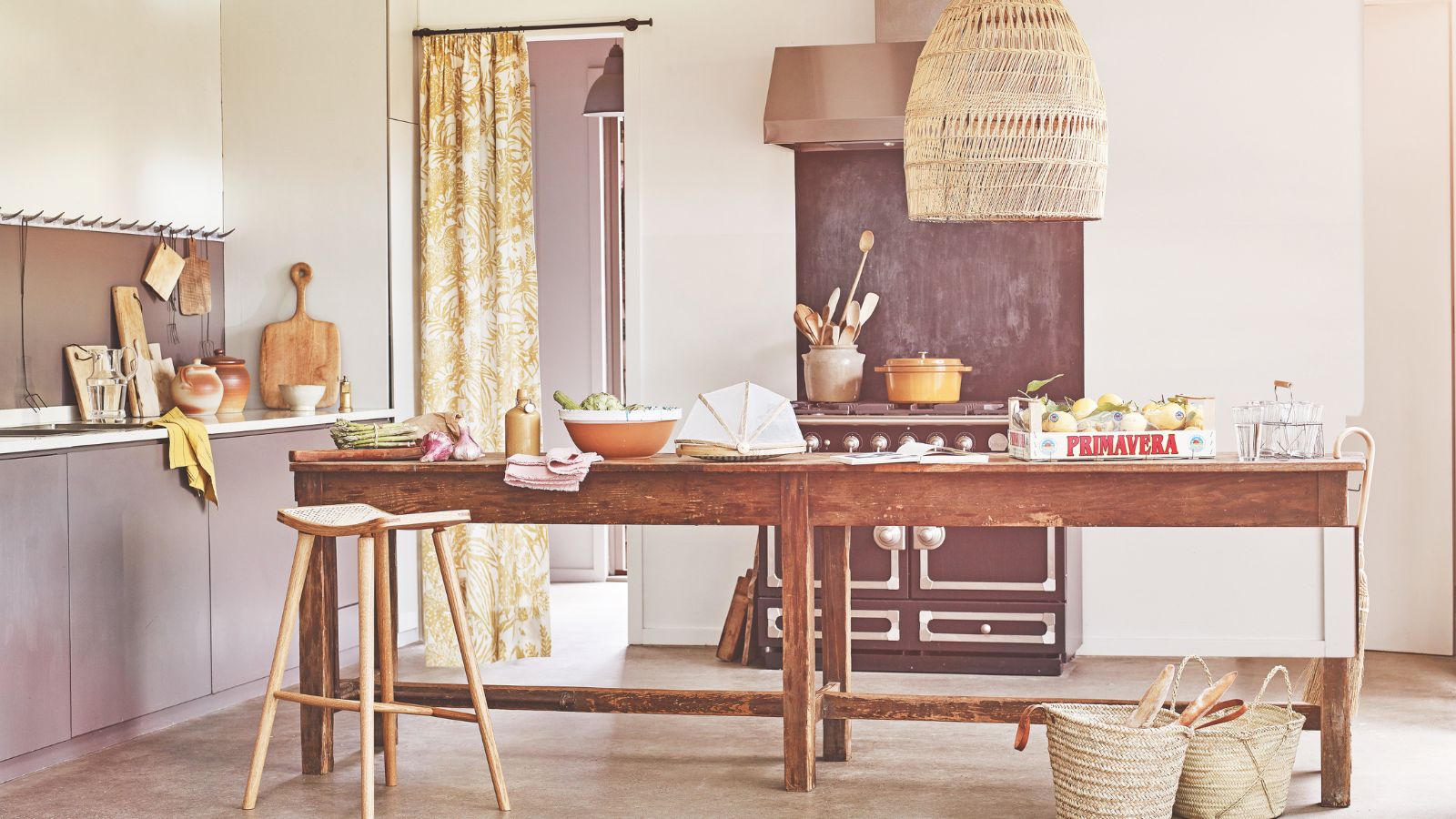 A purple kitchen with a brown wooden table with fruit and bowls, wicker lamp above and rattan baskets. Behind is a purple hob.