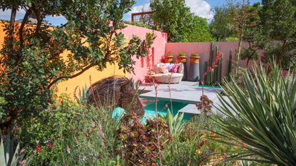 A colorful garden with two walls pink and one wall yellow with a loveseat in the corner in front of the pool and foliage all around
