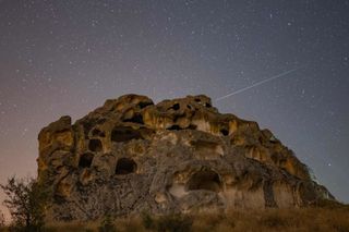 Perseid meteor train across the sky above a large rock structure with numerous carved caves and openings.