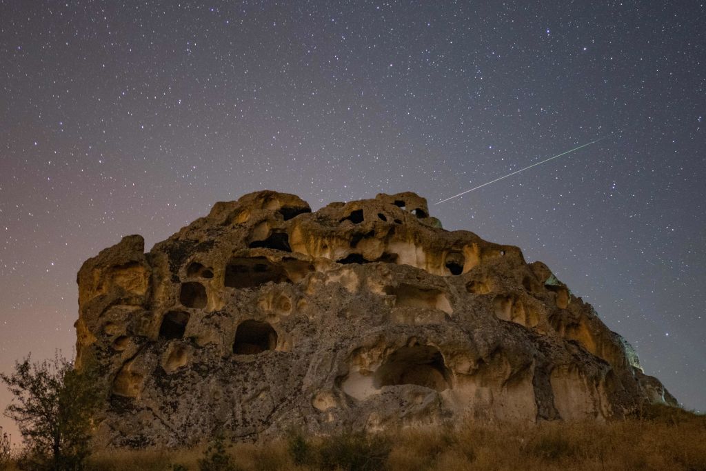 The Perseid meteorite trains in the sky above a large rock formation with many carved caves and openings.