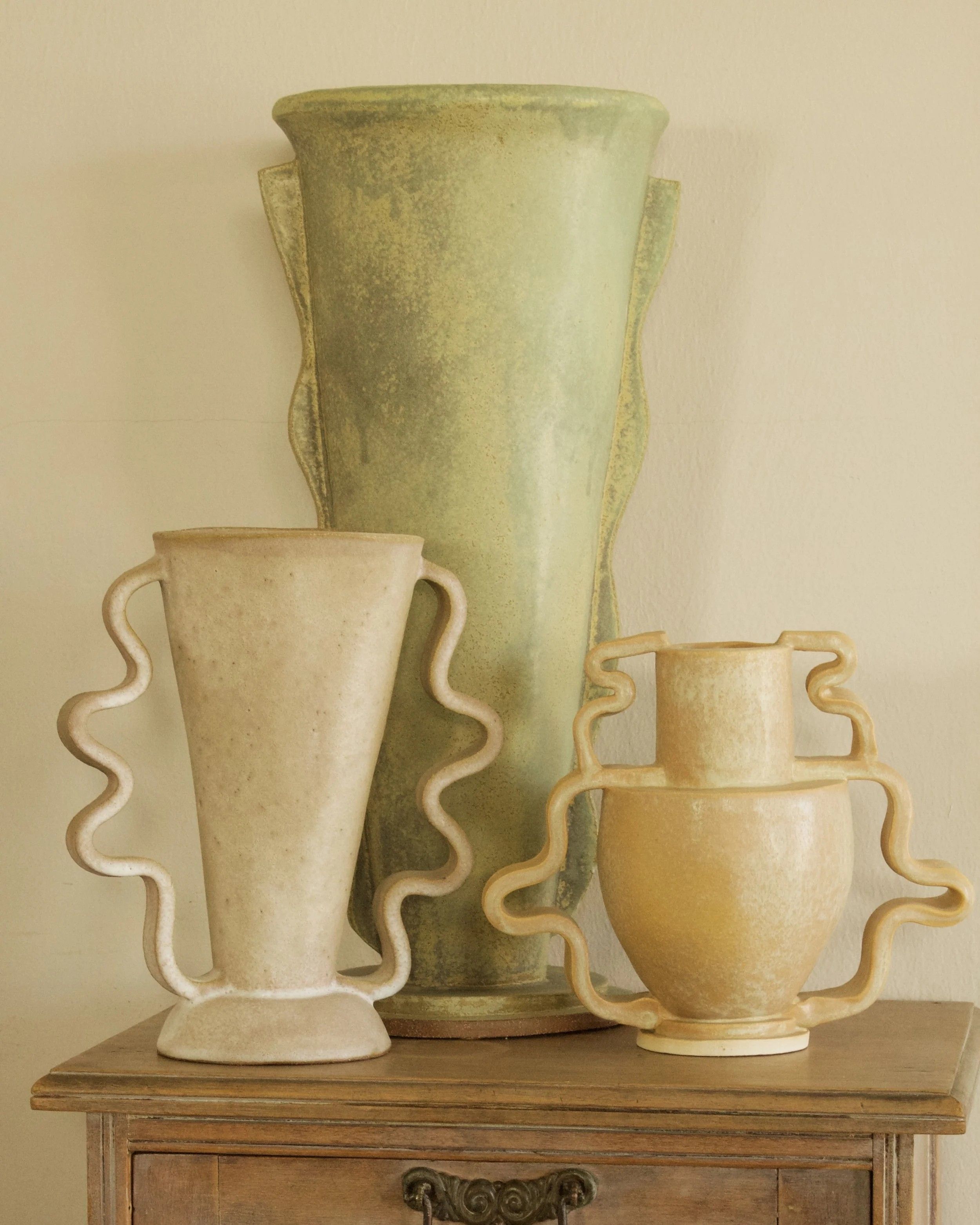 A series of three ceramic vases in white-washed terracotta, pale green, and buttery tones, respectively, sit with their wavy handles on a wooden dresser.