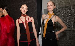 Three female models wearing looks from Prada's collection. One model is wearing a red long hair fur piece and red neck scarf. Another model is wearing a brown neck scarf and black dress with red straps and detail. And the third model is wearing a mustard coloured neck scarf and black dress with silver straps and detail
