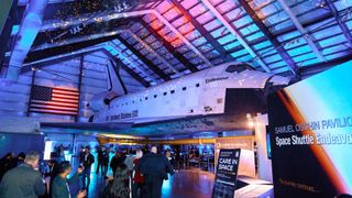 A crowd mingles beneath the sitting Endeavour Space Shuttle. A Care in Space sign sits in the foreground. 
