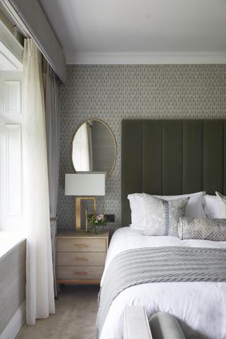 grey bedroom with grey patterned wallpaper, sage green velvet headboard, white bedlinen, gold mirror, gold table lamp, grey curtains