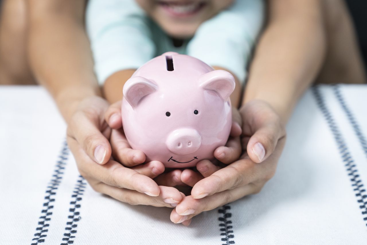 An adult&#039;s hands envelope a child&#039;s hands holding a small piggy bank with a smile on it. 