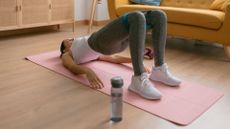 Woman doing glute bridge exercise with resistance band on yoga mat at home, next to sofa