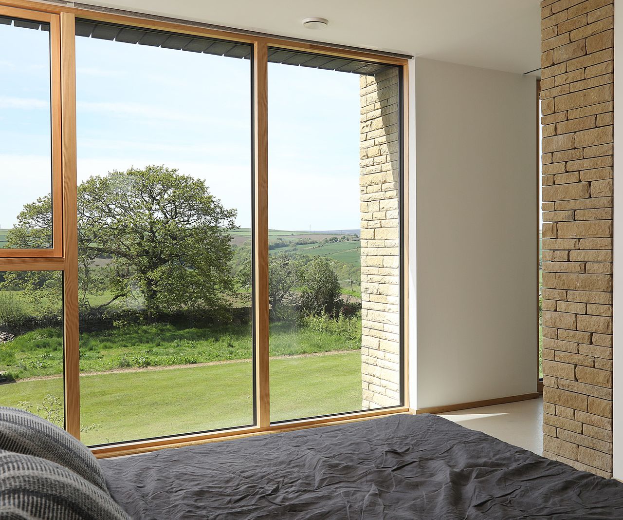 Floor to ceiling glazing across a wall of a bedroom looking out to the surrounding countryside. There are exposed bricks as well as lightly painted walls