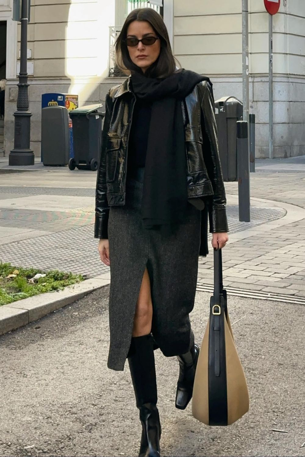 Influencer @leiasfez wears a patent leather jacket with a wool midi pencil skirt and black knee-high leather boots. She accessorises with sunglasses, a dark scarf and a light beige bag.