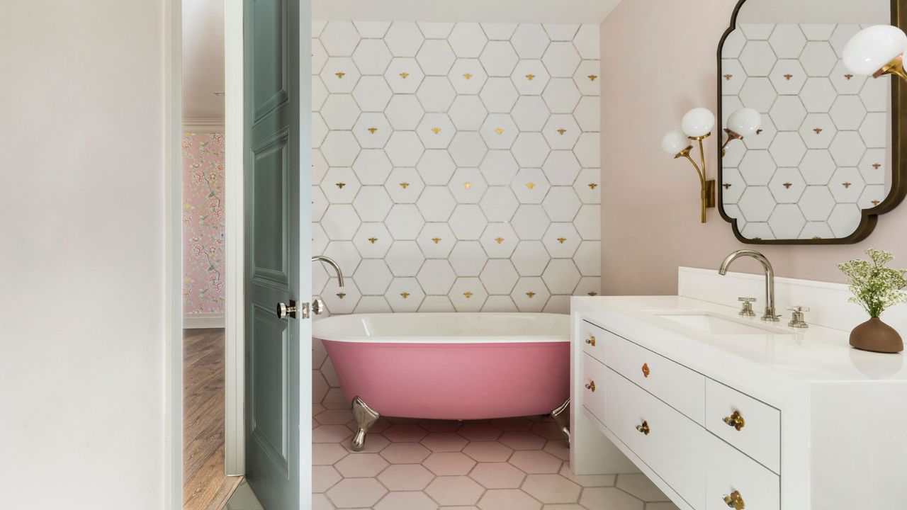 Bathroom with pink painted roll top tub, large hexagon white tiles on one wall with gold bees, matching floor, white vanity, large mirror, globe lights, 