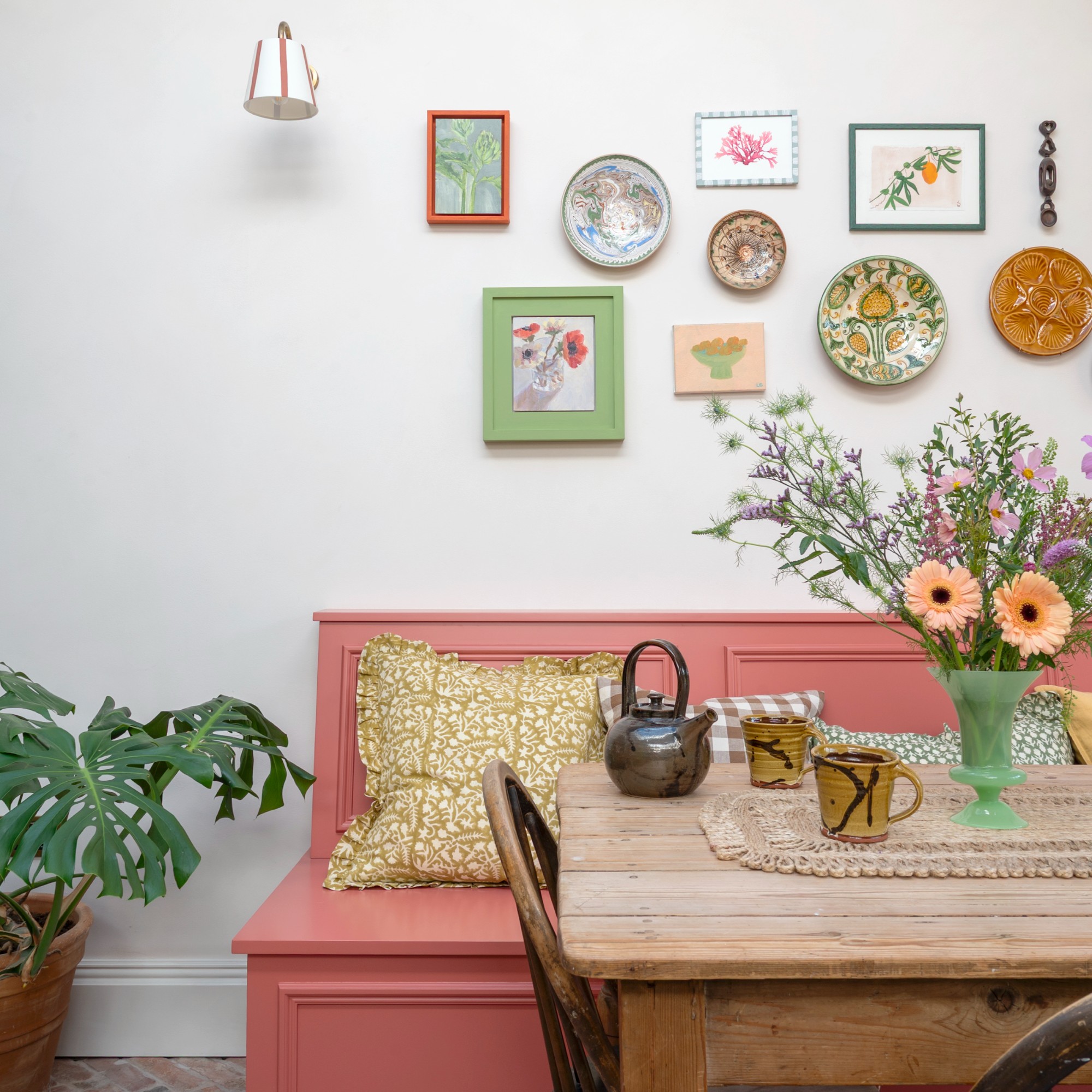 A dining nook with a vintage wooden dining table and a pink-painted dining bench with a gallery wall of framed pictures and decorative plates hanging above